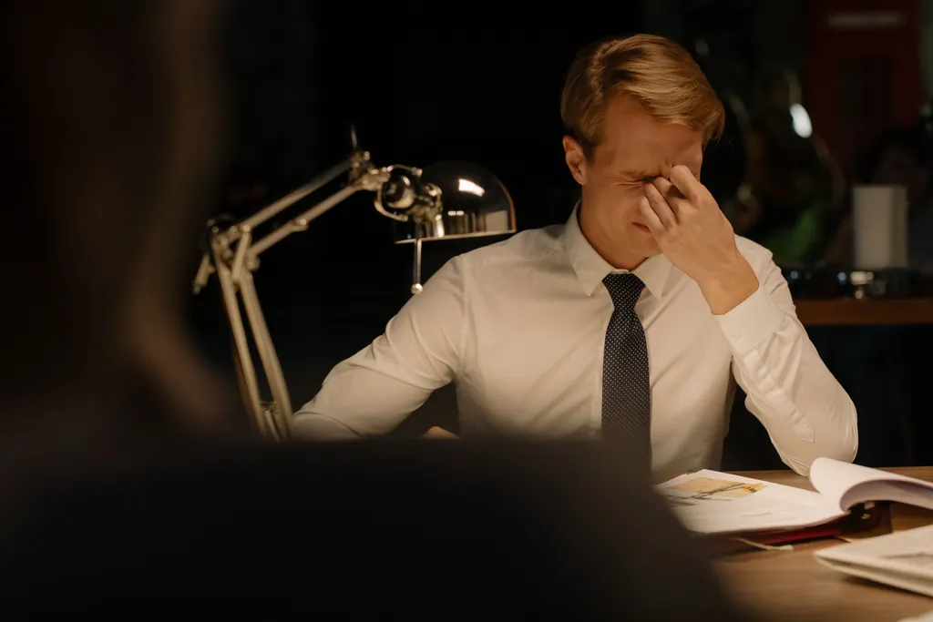 Man in a shirt and tie at a desk pinching the bridge of his nose in frustration