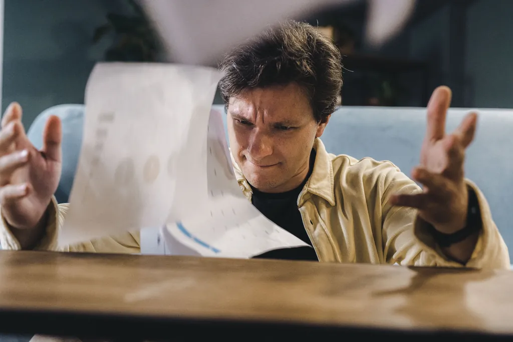 Frustrated young man at a wooden table tossing a sheet of paperwork into the air Frustrated young man at a wooden table tossing a sheet of paperwork into the air